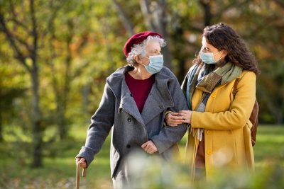 Lovely granddaughter walking with senior woman holding stick in park and wearing mask for safety against covid-19.