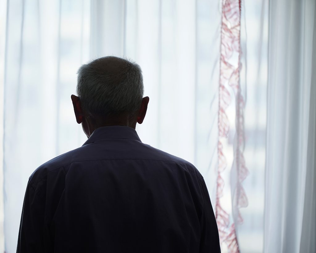 Silhouette of retired man looking through window with transparent curtain